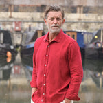 Man wearing a red hemp shirt standing in front of water with boats in the background