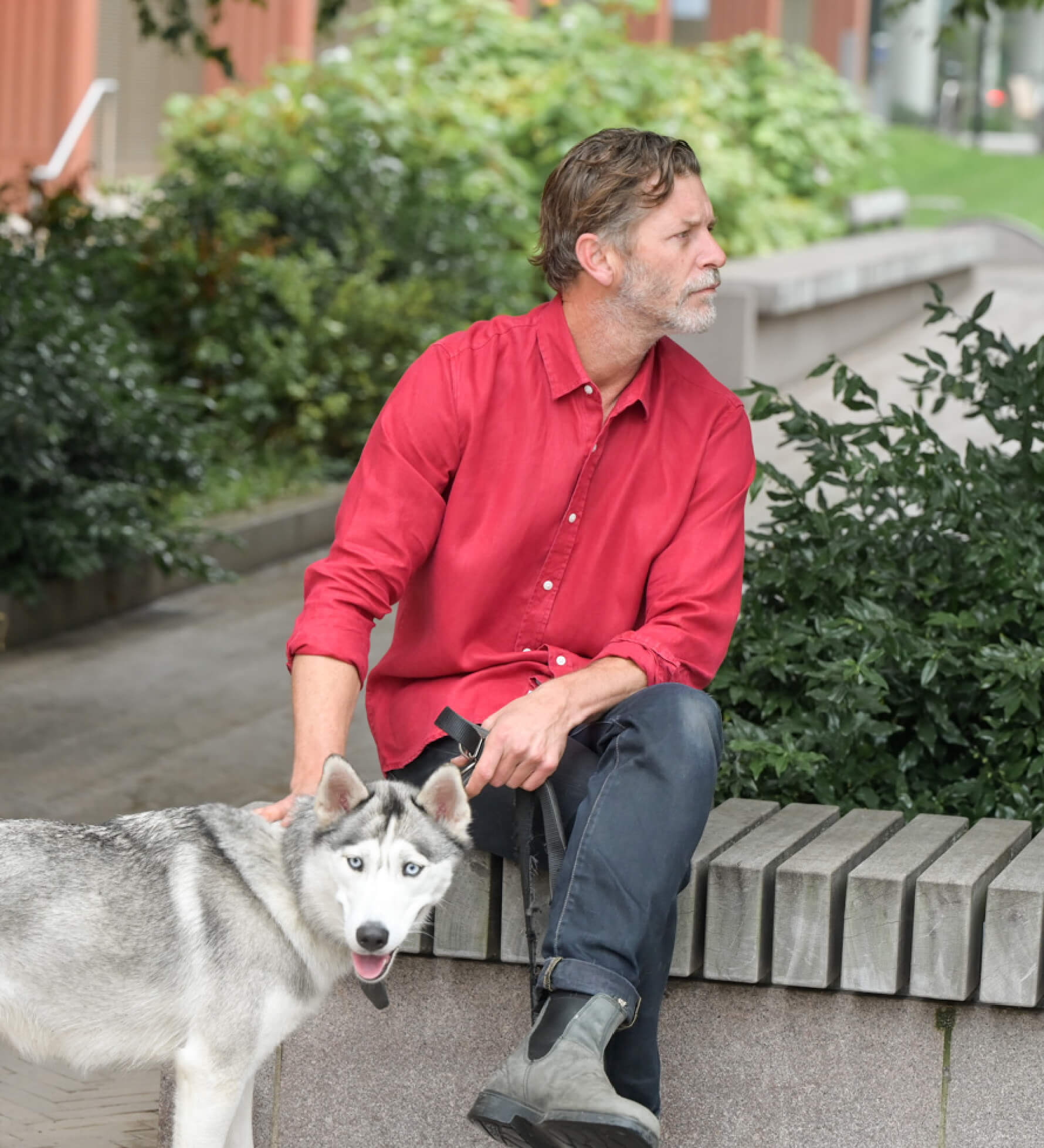 Man in a red shirt sitting on a bench with a dog in an urban setting