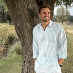 Man in a light blue striped shirt and white pants standing under a tree in a natural setting
