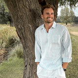 Man in a light blue striped shirt and white pants standing under a tree in a natural setting