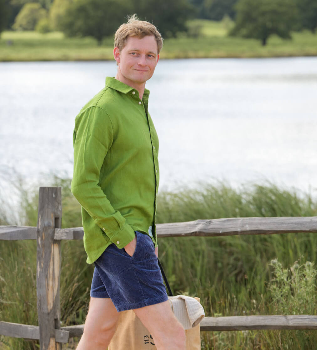 Man in a green shirt and blue shorts standing by a wooden fence with a lake and trees in the background.