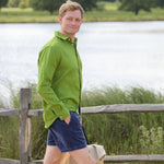 Man in a green shirt and blue shorts standing by a wooden fence with a lake and trees in the background.