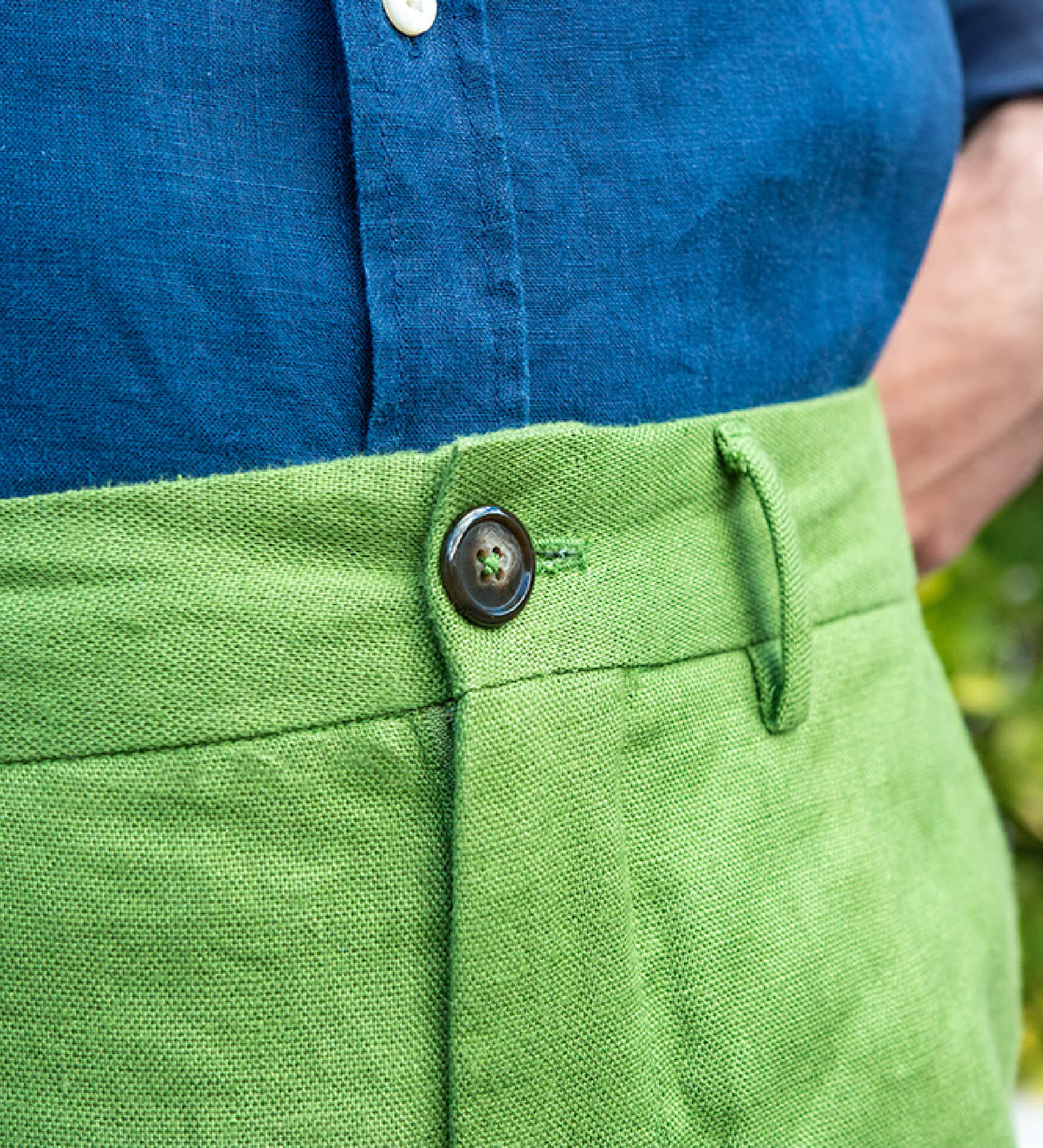 Close-up of green hemp shorts with a button, worn with a blue shirt against a blurred natural background.