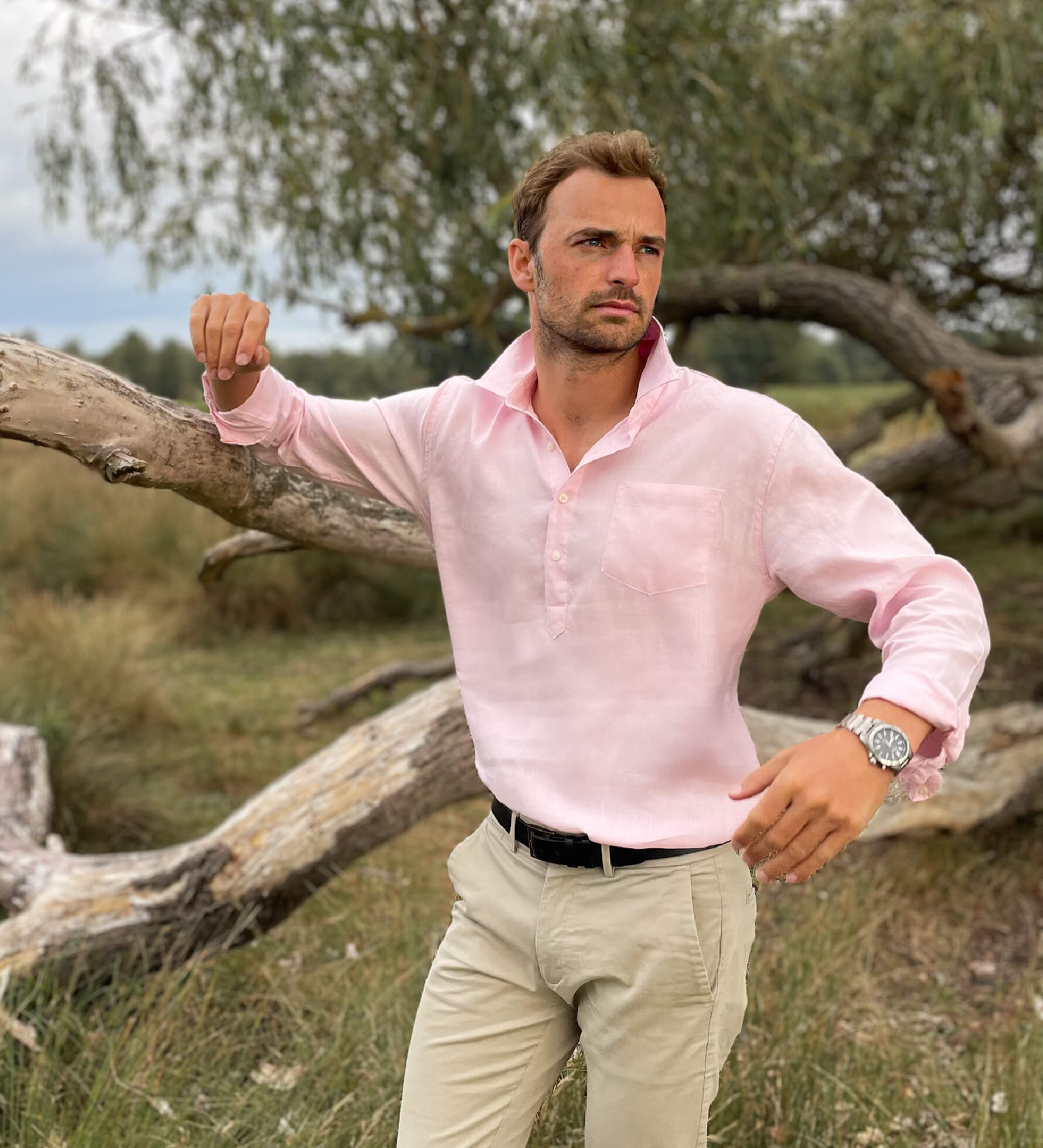 Man wearing a pink hemp shirt and beige pants standing outdoors leaning on a tree with nature in the background