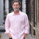 Man wearing a light pink shirt standing outdoors near a building. His hands are in his pocket and he smiles at the camera, the shirt is made from soft hemp fabric.