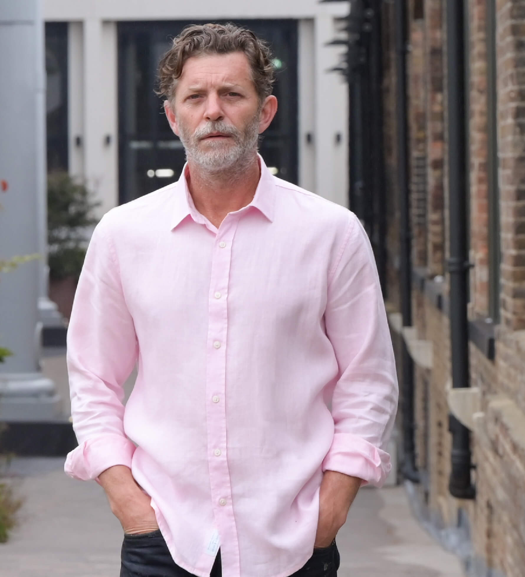 Man wearing a light pink shirt standing outdoors near a building. His hands are in his pocket and he smiles at the camera, the shirt is made from soft hemp fabric.
