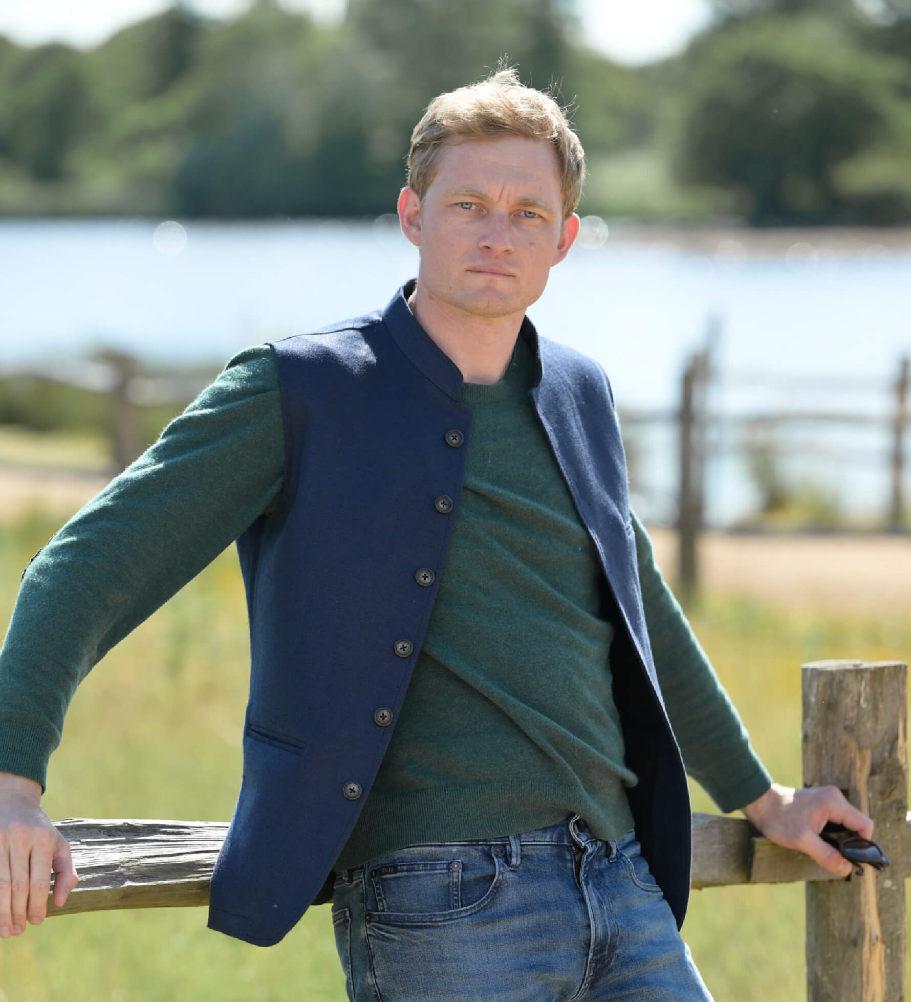 Man standing by a wooden fence with a scenic background of trees and water.