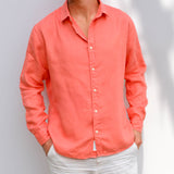 Person wearing a coral button-up shirt against a plain background. Both arms have sleeves rolled down and hands in pockets.