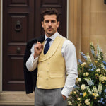man in yellow hemp waistcoat with shirt and tie on stands in front of doorway to large house