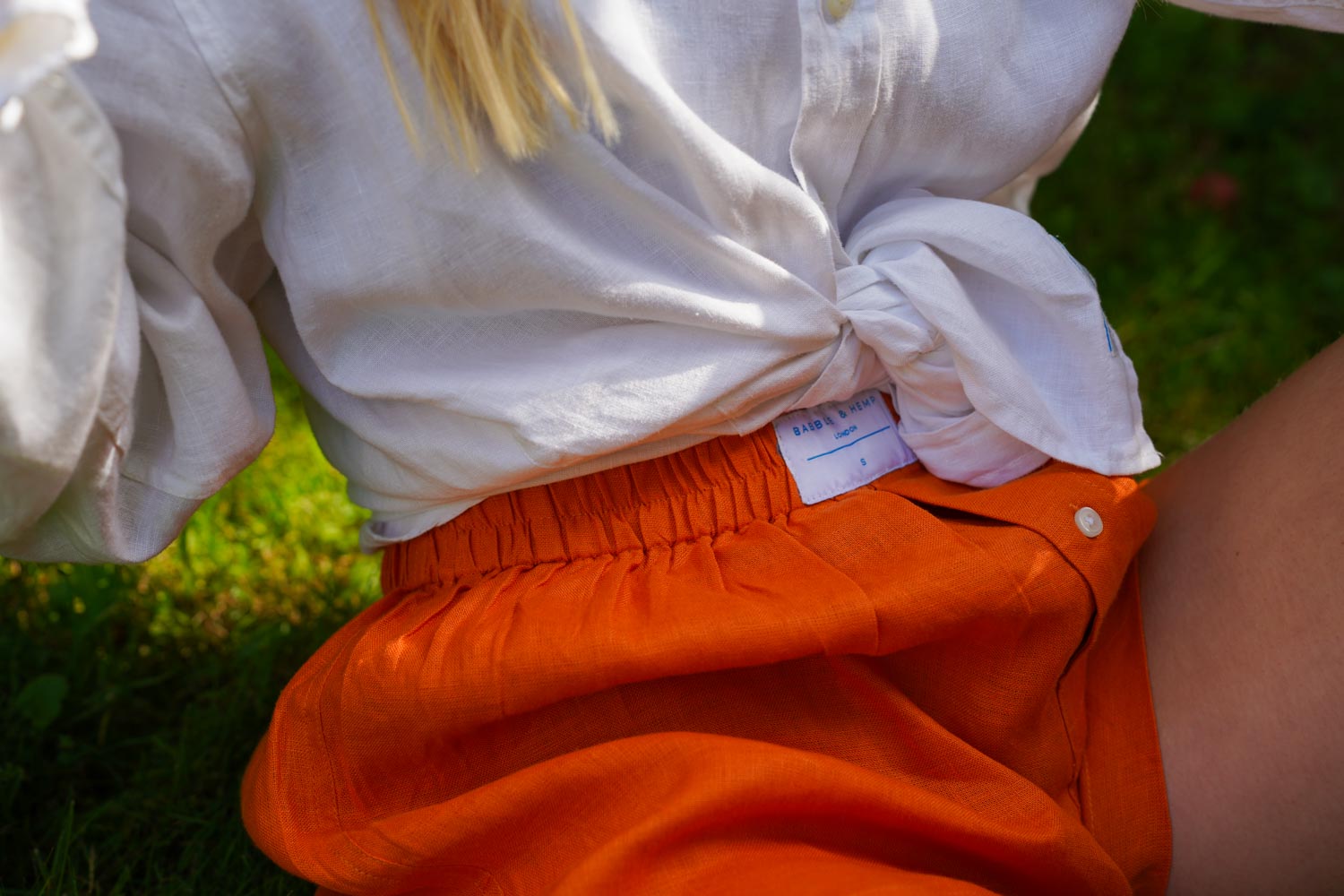 Person wearing a white shirt and orange hemp boxer shorts with a visible brand Babble & Hemp label, sitting on grass.