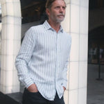 a video showing a man leaning next to a pillar with hands in pocket whilst wearing a blue and white stripe hemp shirt and dark blue trousers