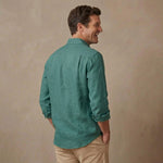 A man wearing a teal green hemp long-sleeve shirt and beige pants, shown from a side-back angle against a textured beige studio backdrop.