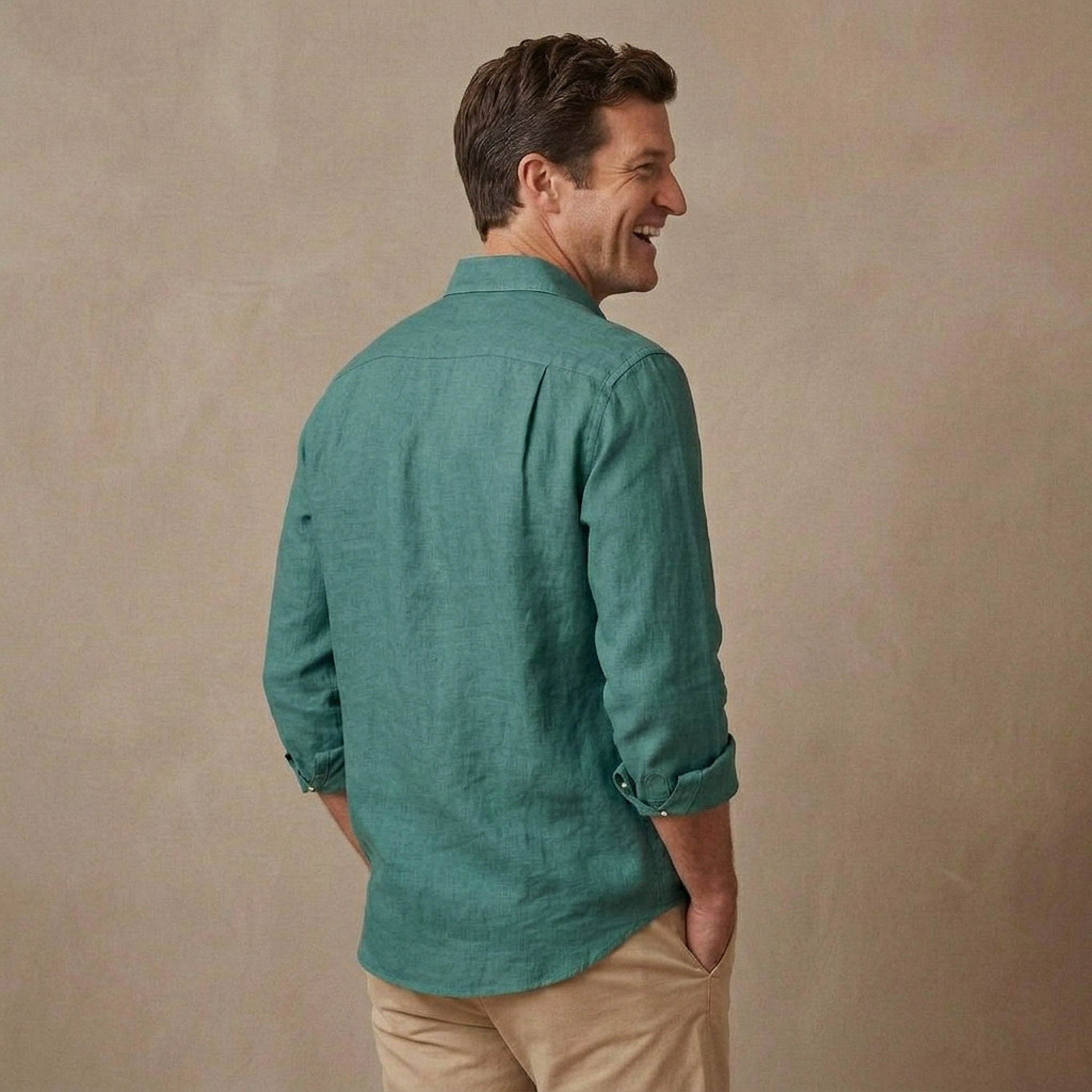 A man wearing a teal green hemp long-sleeve shirt and beige pants, shown from a side-back angle against a textured beige studio backdrop.