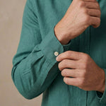 Close-up of a man's hands adjusting the button on the cuff of a teal green hemp  shirt against a neutral beige background.