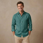 A smiling man wearing a teal-green long-sleeve hemp shirt with beige pants, standing against a textured beige studio background.