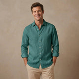 A smiling man wearing a teal-green long-sleeve hemp shirt with beige pants, standing against a textured beige studio background.
