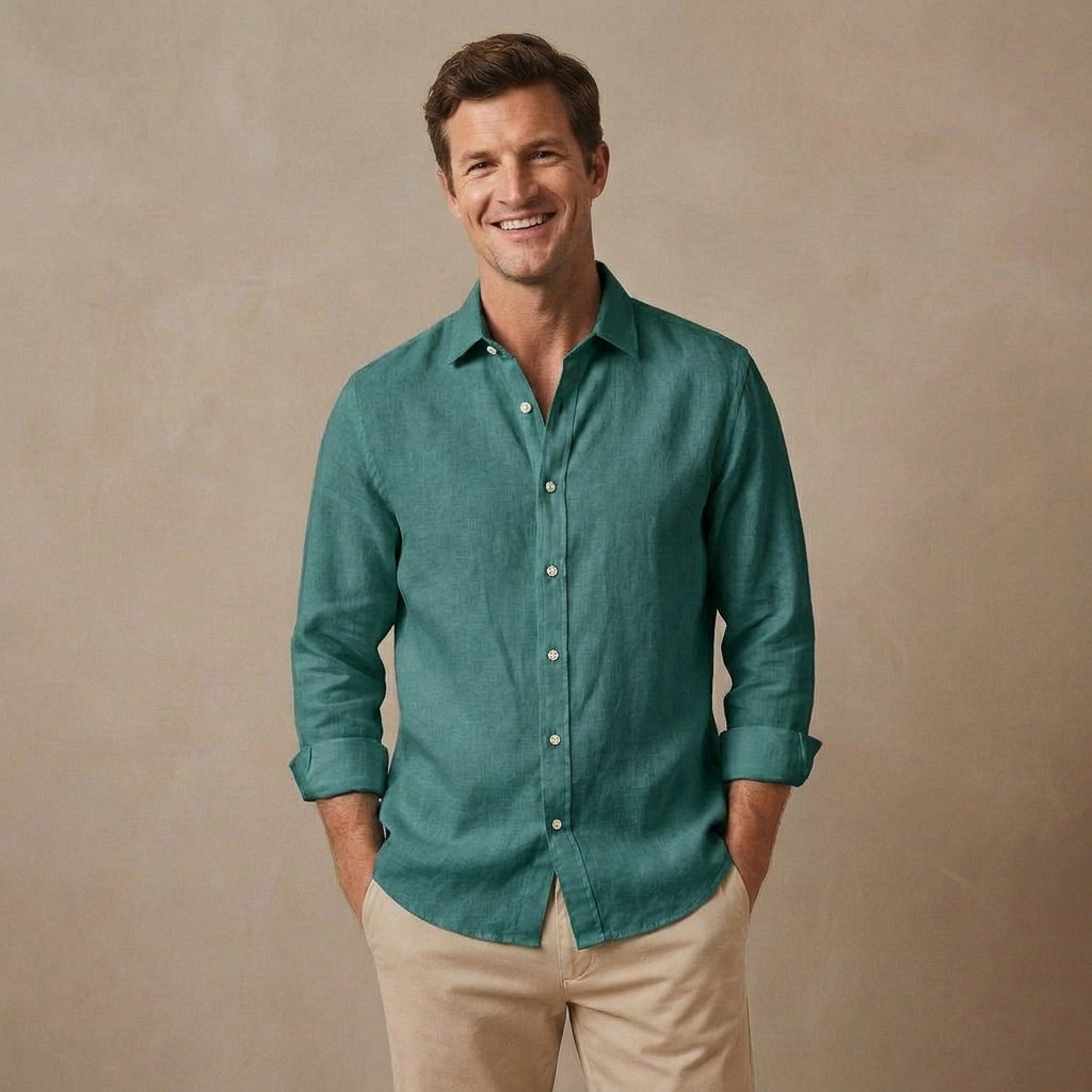 A smiling man wearing a teal-green long-sleeve hemp shirt with beige pants, standing against a textured beige studio background.