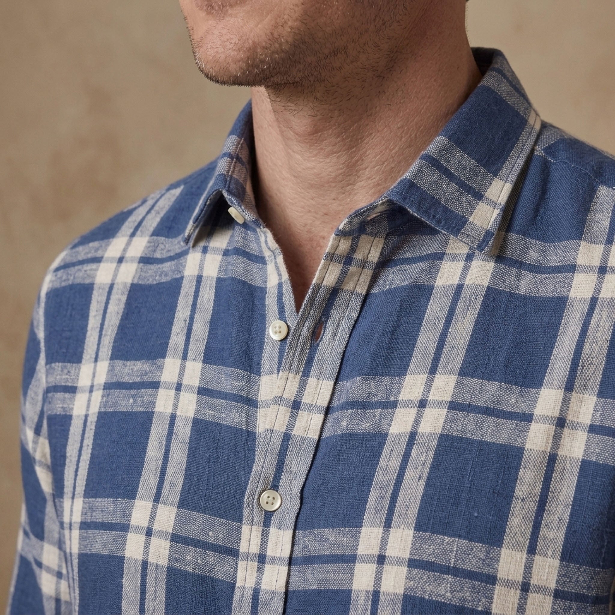 Close-up of a man wearing a blue and off-white plaid heavy hemp shirt, showing the collar, chest, and part of the neck against a neutral beige background.