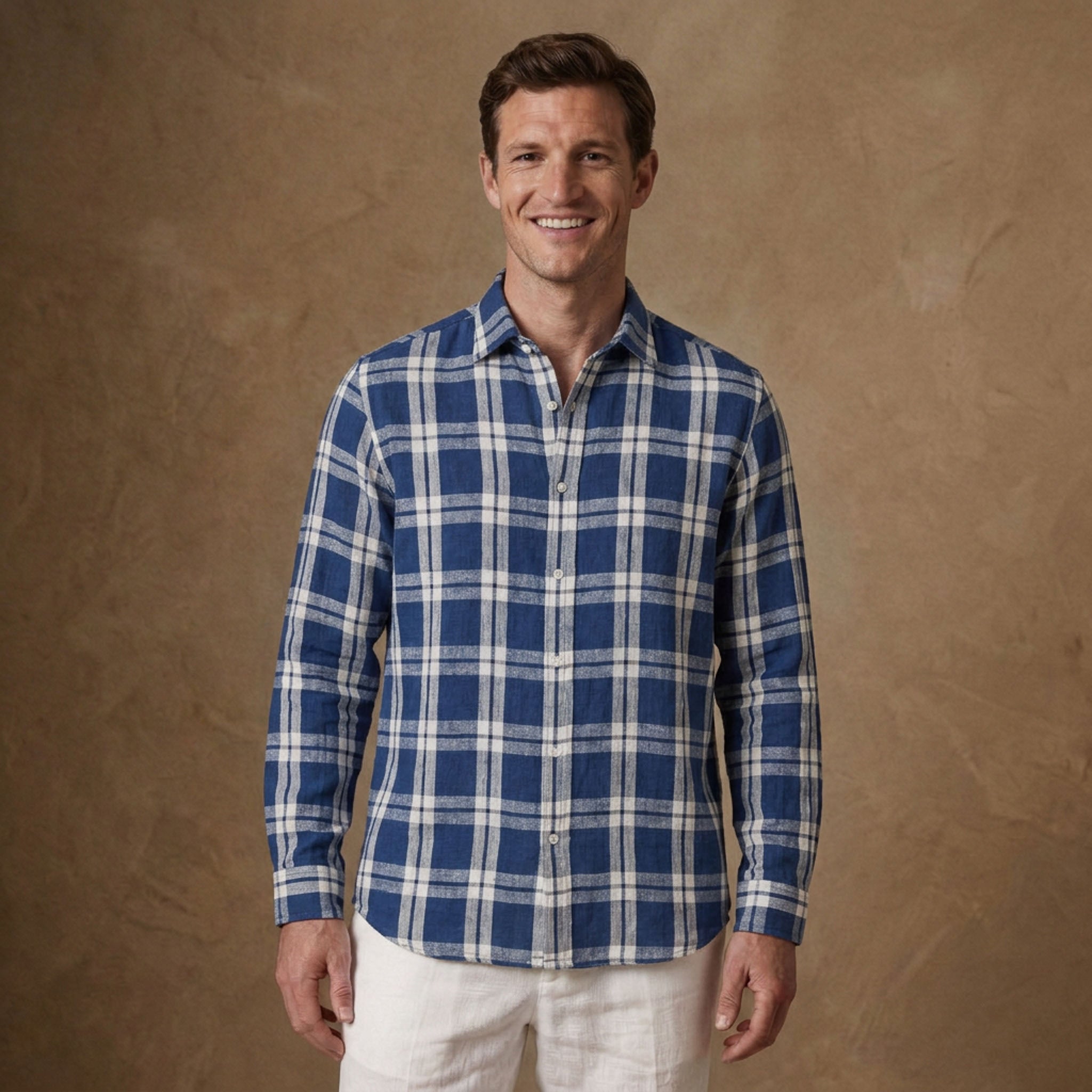 A man wearing a blue and white plaid long-sleeve button-down soft hemp shirt, paired with white pants, standing against a textured brown studio backdrop.