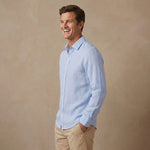 A man wearing a light blue long-sleeve soft hemp shirt and khaki pants, standing against a textured beige studio background.