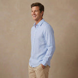 A man wearing a light blue long-sleeve soft hemp shirt and khaki pants, standing against a textured beige studio background.