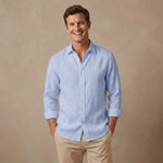 A smiling man wearing a light blue long-sleeve button-up  soft hemp shirt with beige pants, standing against a warm beige studio background.