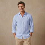 A smiling man wearing a light blue long-sleeve button-up  soft hemp shirt with beige pants, standing against a warm beige studio background.