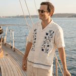 a photo of a man wearing a smart casual, camp collared hemp shirt made from cream or off white coloured hemp fabric with floral embroidery whilst standing on a yacht
