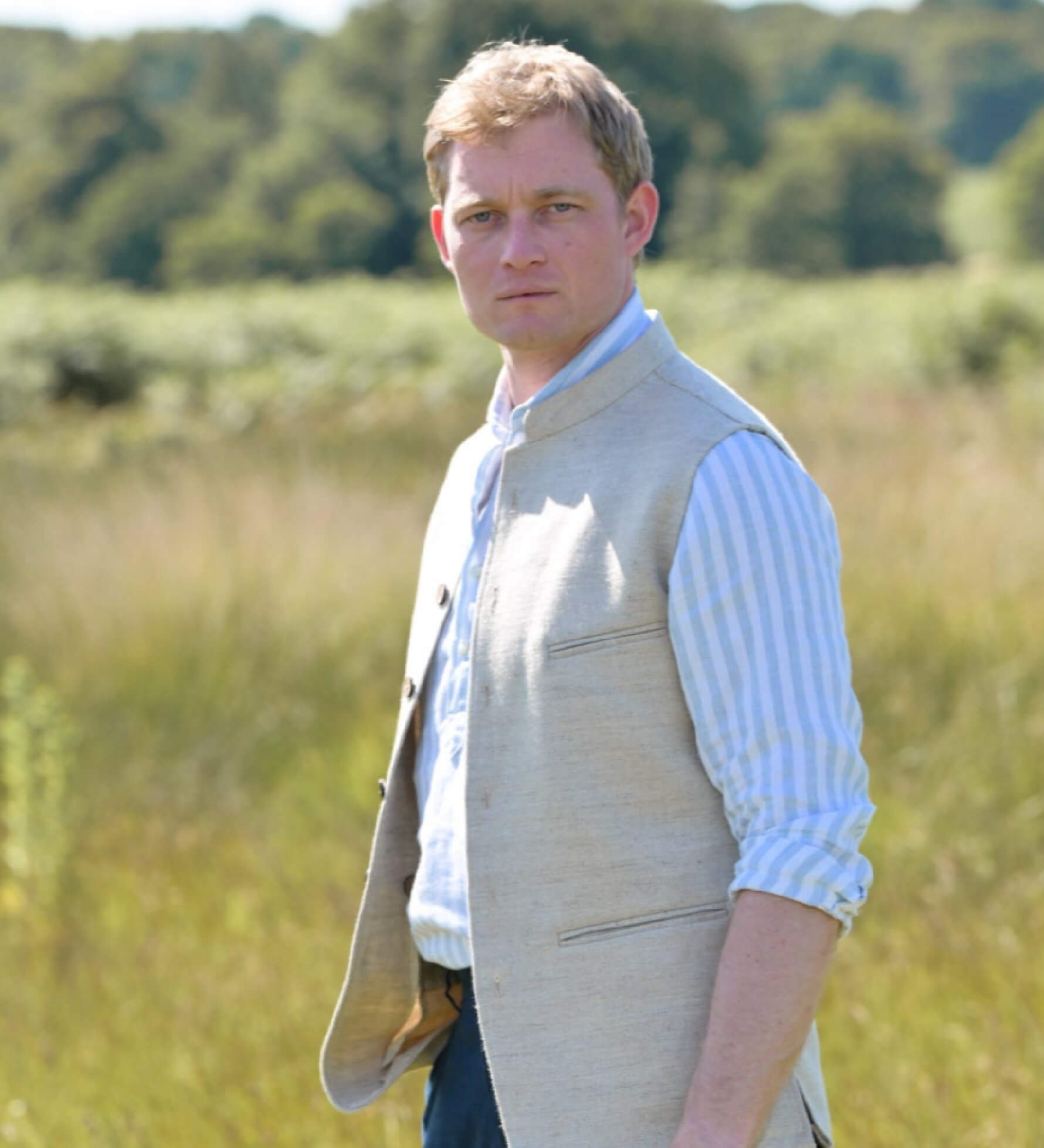 Man wearing a beige coloured sleeveless nehru jacket over a light blue shirt in a field with trees in the background