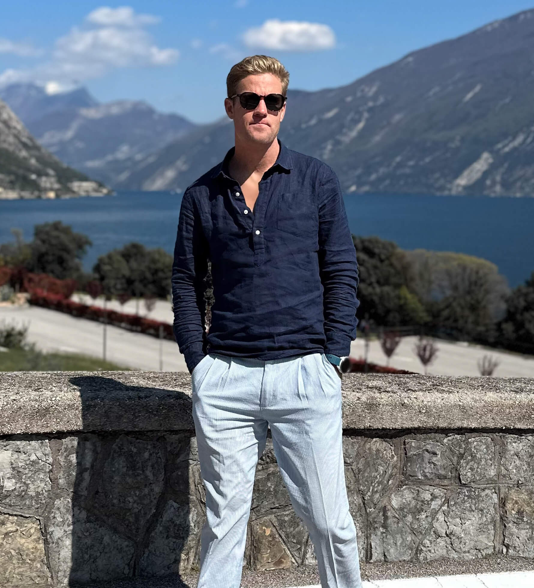 Man in sunglasses and a navy blue hemp shirt standing against a scenic backdrop of mountains and water.