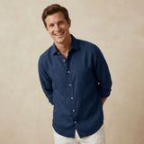 Man wearing a navy blue hemp shirt and white trousers against a beige background whilst laughing and smiling