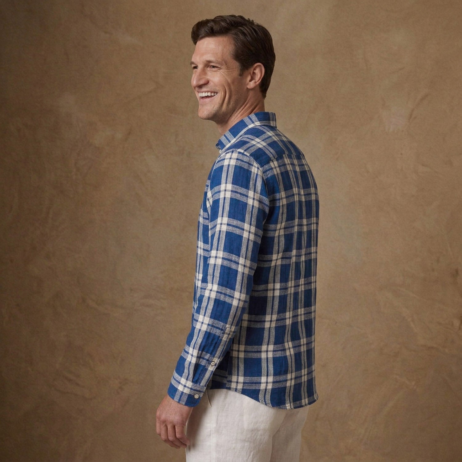 A man wearing a blue and white plaid long-sleeve button-down shirt, shown from a side-back angle against a textured brown studio backdrop.