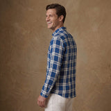 A man wearing a blue and white plaid long-sleeve button-down shirt, shown from a side-back angle against a textured brown studio backdrop.