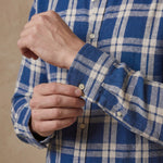 Close-up of a person wearing a blue and off-white plaid long-sleeve hemp shirt, adjusting the cuff with one hand while the other hand fastens a button.