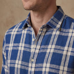 Close-up of a man wearing a blue and off-white plaid button-down shirt, showing the collar, chest area, and part of the neck against a neutral beige background.