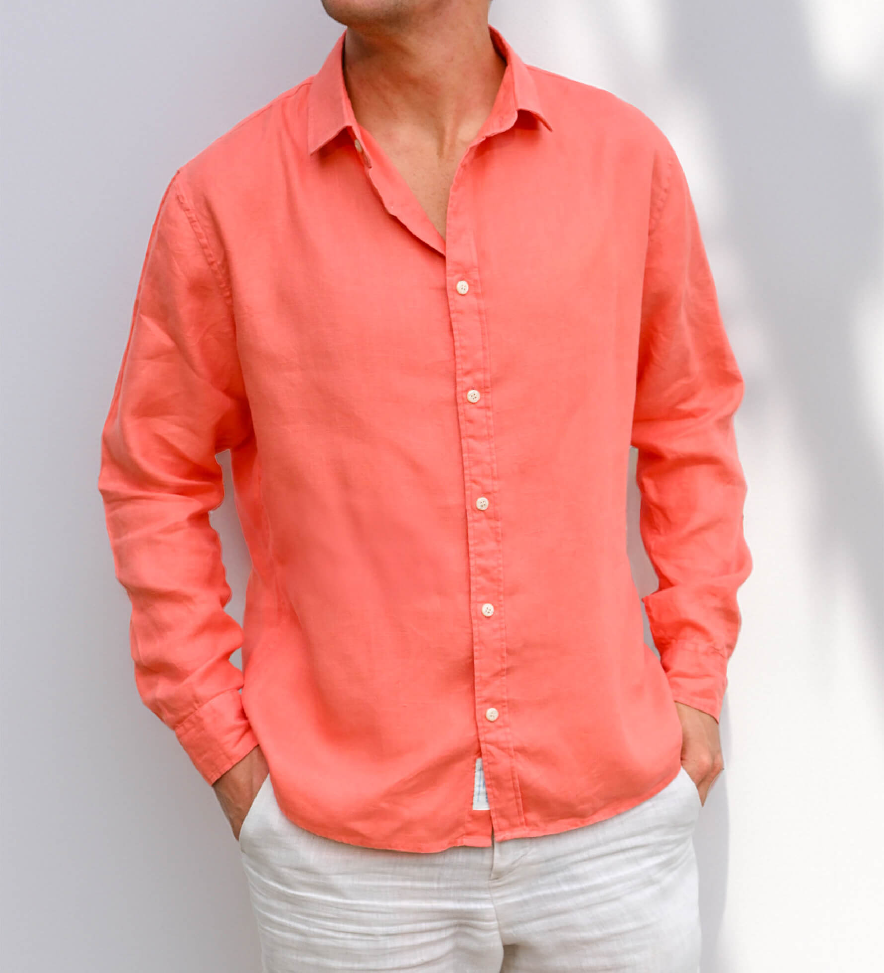 Person wearing a coral button-up shirt against a plain background. Both arms have sleeves rolled down and hands in pockets. 
