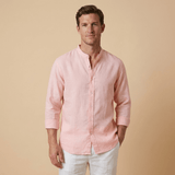 Man wearing a light pink or peach coloured hemp shirt and white pants against a beige background. the shirt has a grandad collar