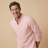 Man wearing a light pink or peach coloured hemp shirt and white pants against a beige background. the shirt has a grandad collar and his sleeves are rolled up and he smiles at the camera whilst leaning in.