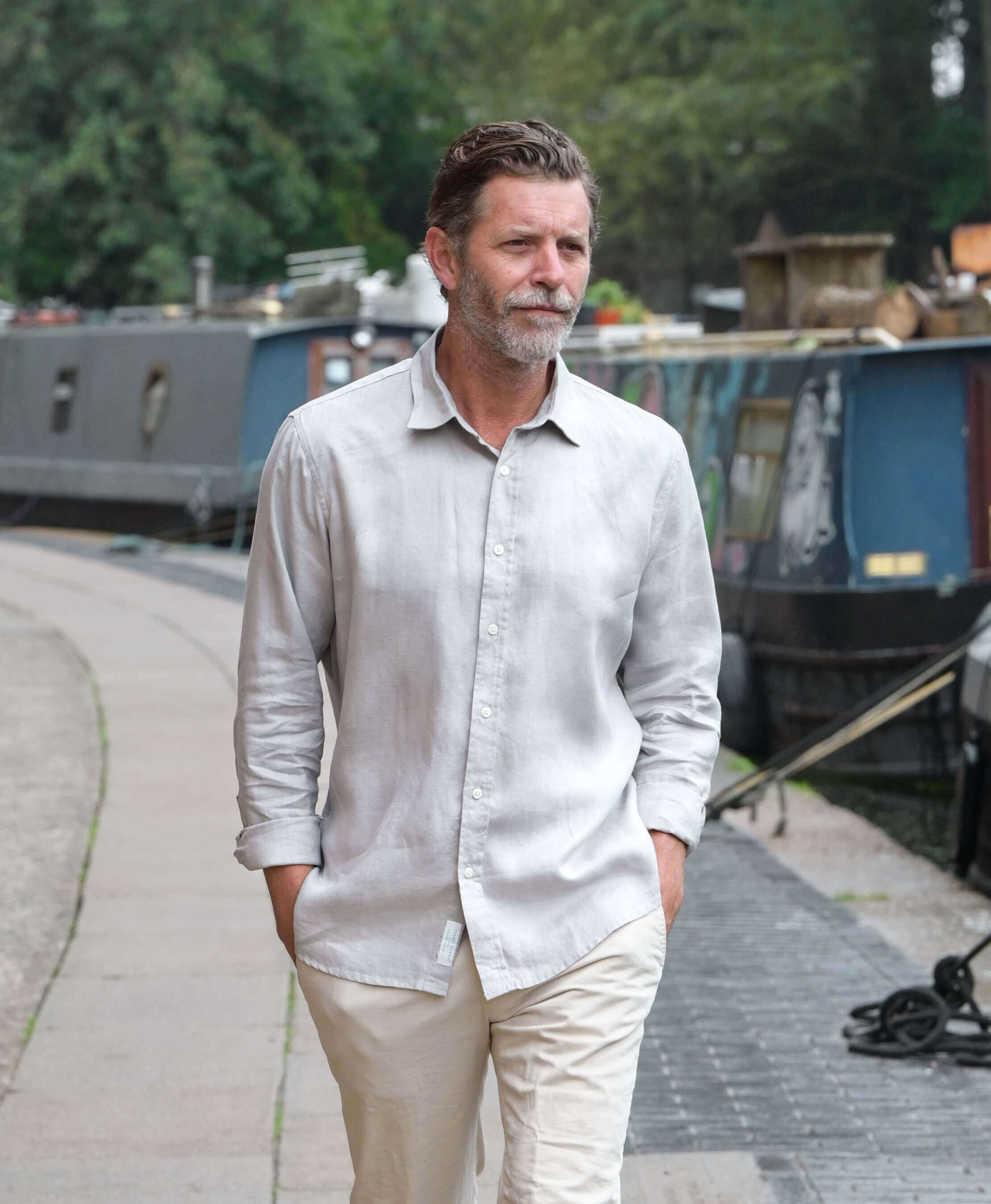 Man in a light grey shirt and beige pants standing on a path with boats in the background. The shirt is made from eco-friendly hemp fabric