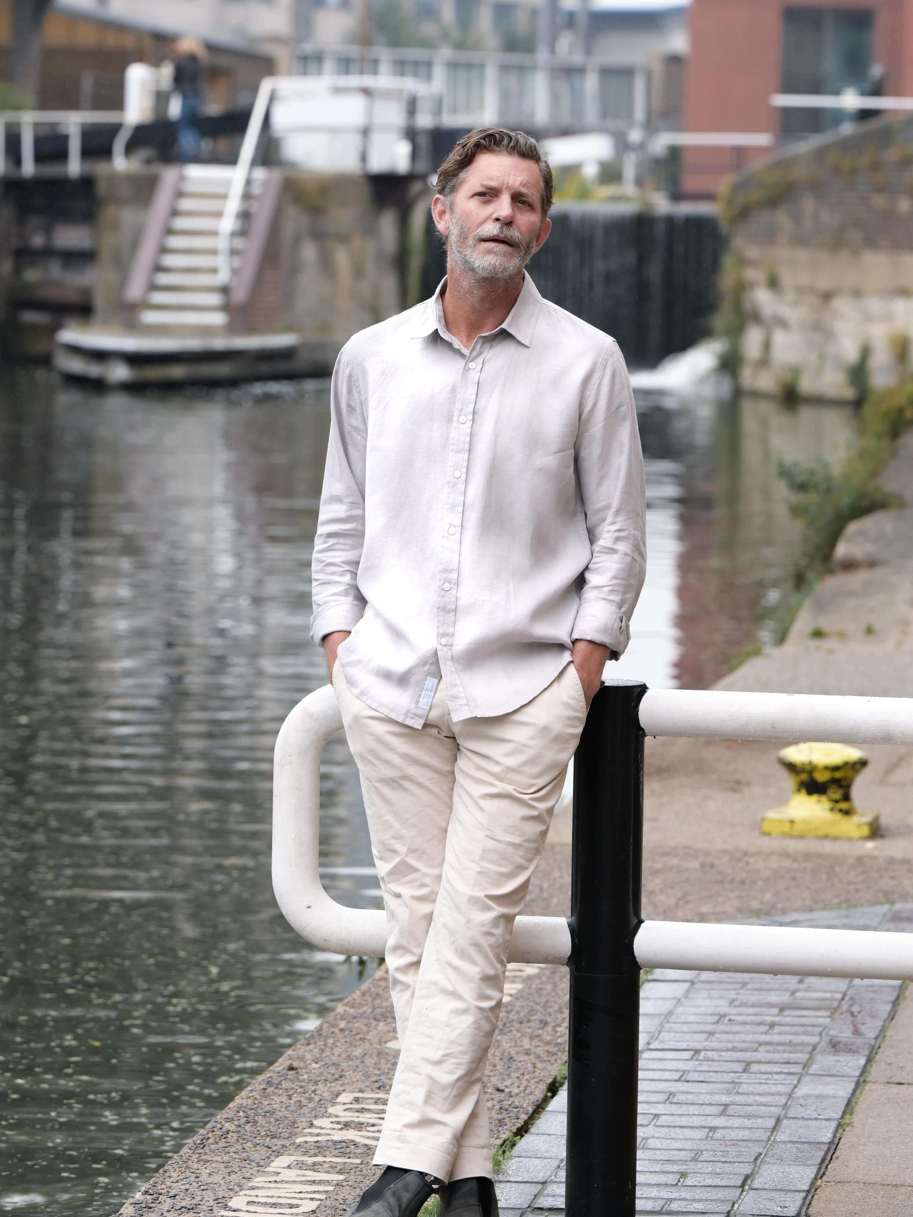 Man in light-colored shirt and pants standing by a canal with buildings in the background