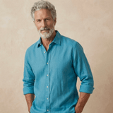 a man models a petrol blue shirt made from hemp fabric which is better than linen set against a beige wall behind him