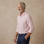 A smiling older man with gray hair and a beard, wearing a light pink and white gingham long-sleeve hempshirt and navy blue trousers, standing against a textured beige studio backdrop.