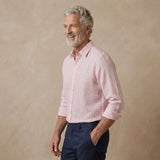 A smiling older man with gray hair and a beard, wearing a light pink and white gingham long-sleeve hempshirt and navy blue trousers, standing against a textured beige studio backdrop.