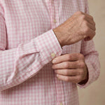 A close-up of a man's hands adjusting the cuff of a light pink and white gingham hemp shirt.