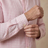 A close-up of a man's hands adjusting the cuff of a light pink and white gingham hemp shirt.