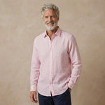A smiling older man with gray hair and a beard wearing a light pink and white gingham long-sleeve soft hemp shirt, paired with dark navy pants, standing against a textured beige studio backdrop.