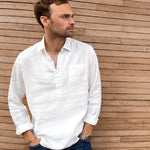 Man wearing a white hemp shirt with buttons only half way down the shirt, in a popover style and jeans against a wooden wall