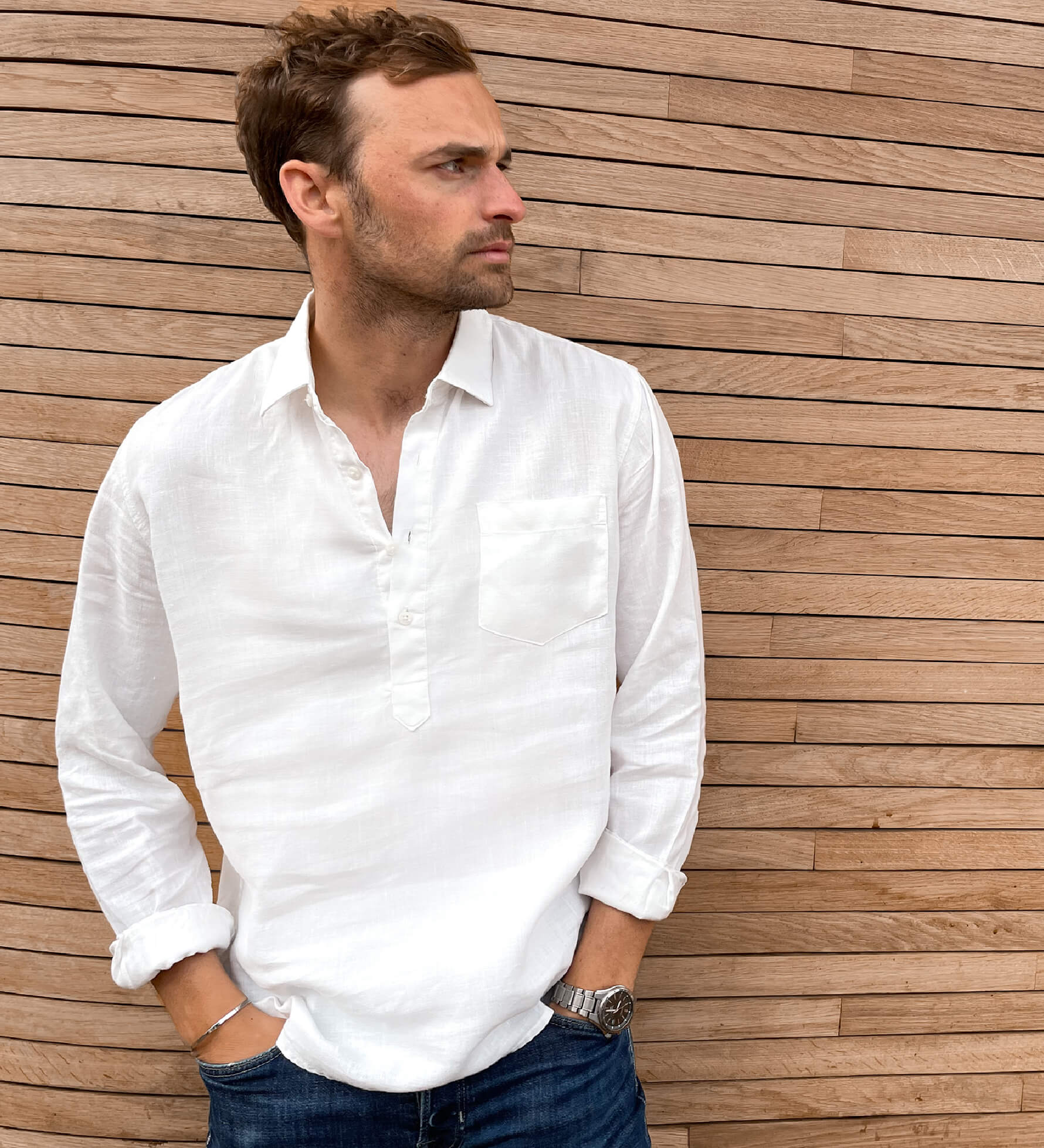 Man wearing a white hemp shirt with buttons only half way down the shirt, in a popover style and jeans against a wooden wall