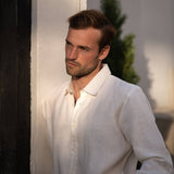 Man wearing a white hemp shirt leaning against a wall with a plant in the background