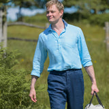 Man in a light blue hemp fabric shirt and dark pants walking outdoors with greenery in the background